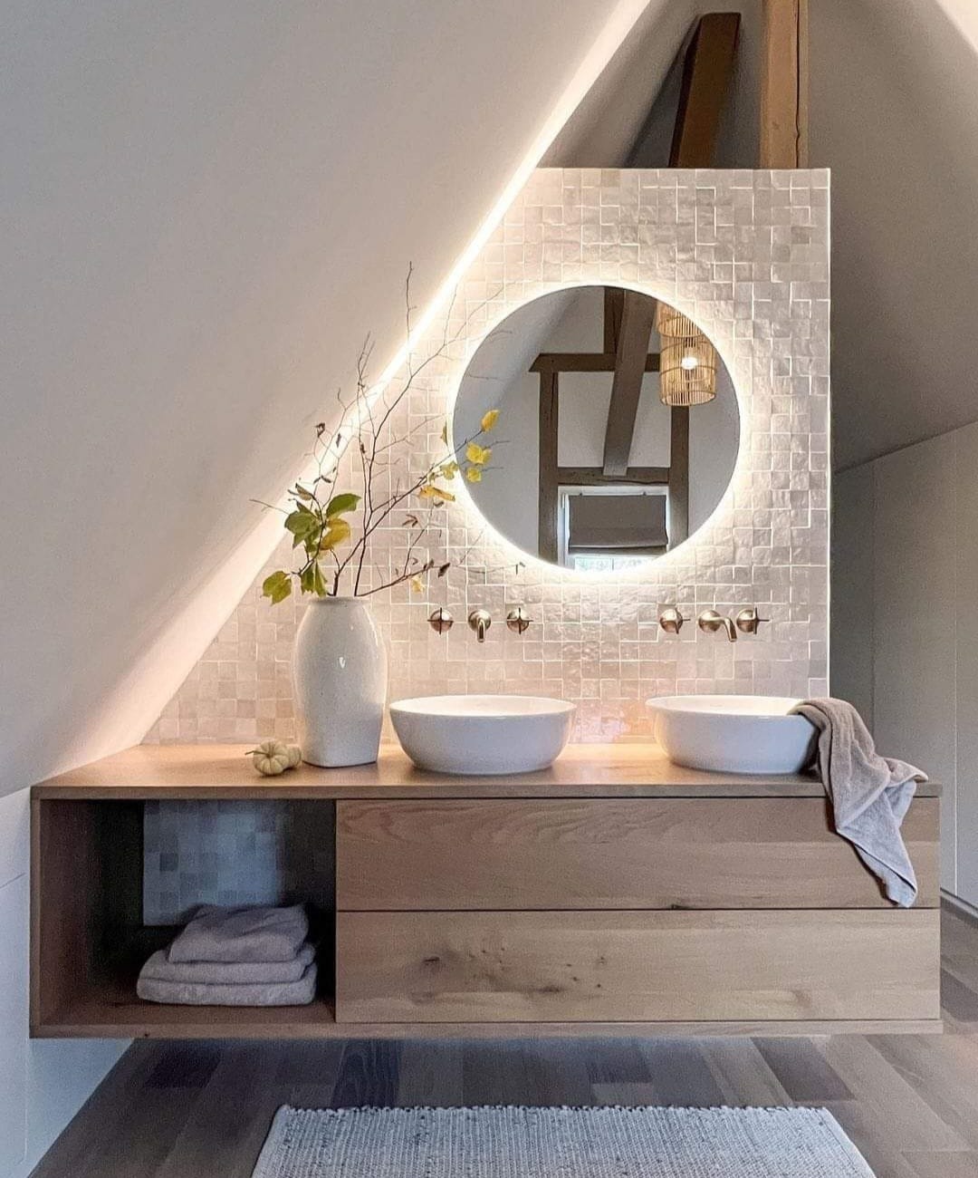Nowoczesna łazienka na poddaszu z dwoma białymi umywalkami nablatowymi, drewnianą szafką z szufladami, okrągłym lustrem z podświetleniem LED i mozaiką na ścianie. W wazonie gałązka z żółtymi liśćmi...