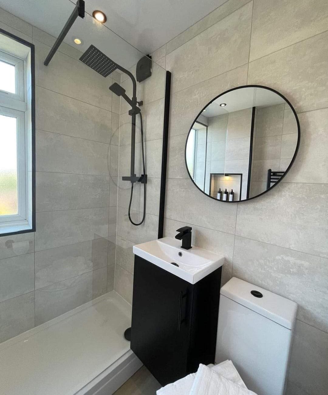 Nowoczesna łazienka z kabiną prysznicową typu walk-in, czarną armaturą, okrągłym lustrem i umywalką z szafką w kolorze czarnym.