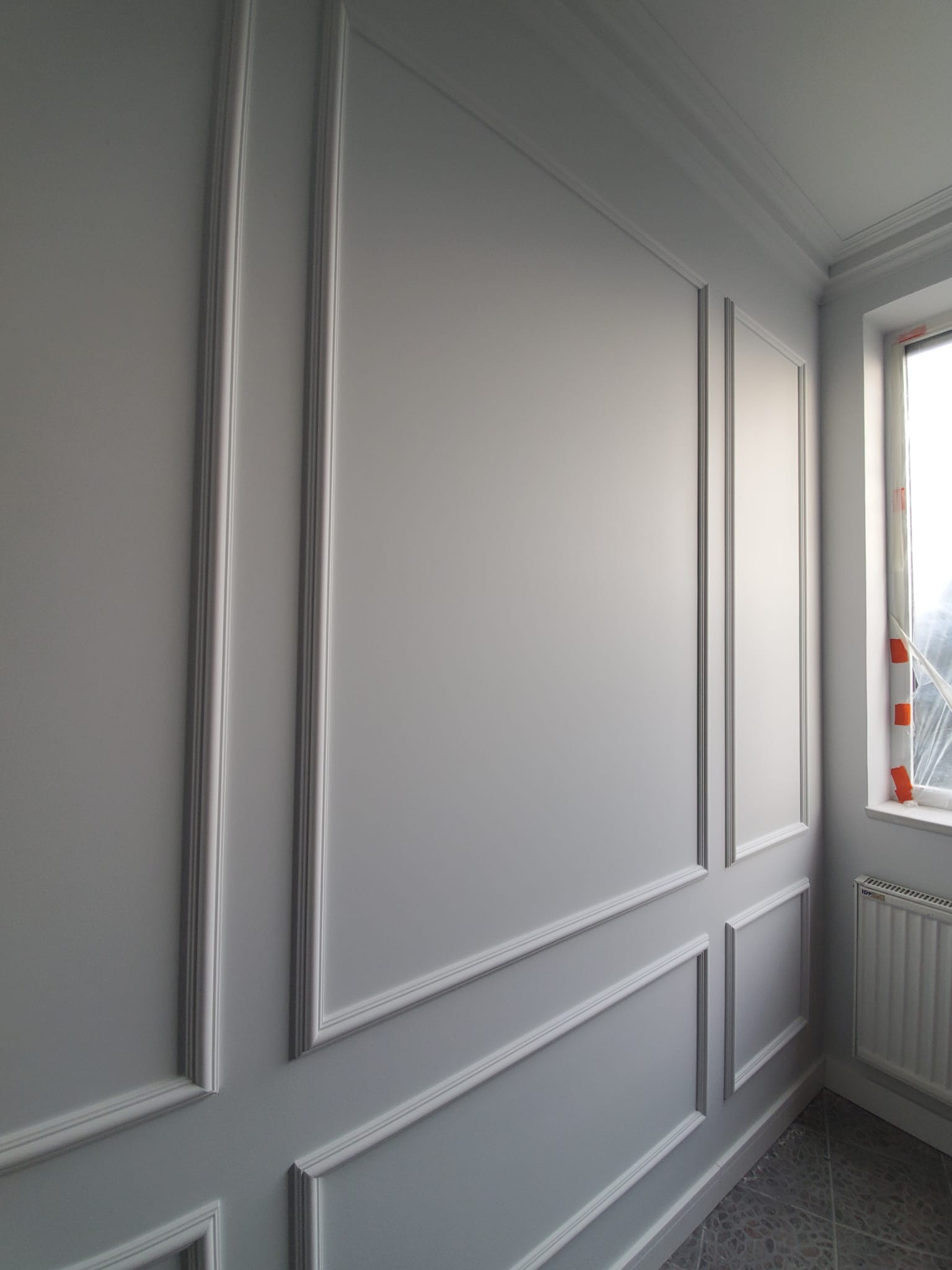 Szara ściana z dekoracyjnymi listwami w stylu klasycznym, widoczne okno z zabezpieczeniem folią malarską i grzejnik.