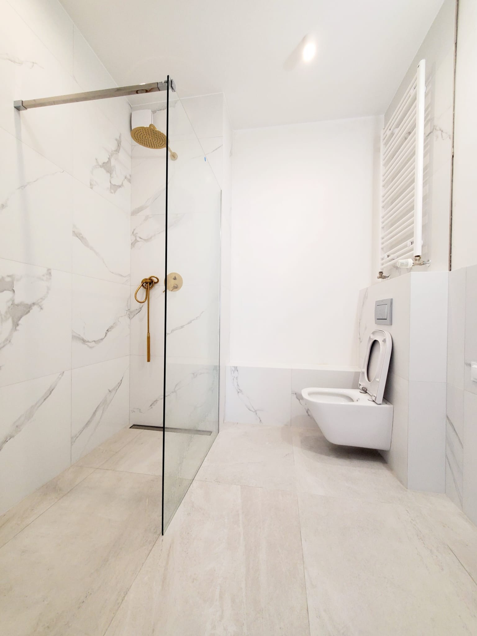 Nowoczesna, jasna łazienka z marmurowymi płytkami, złotą armaturą prysznicową i podwieszaną toaletą z otwartą deską.