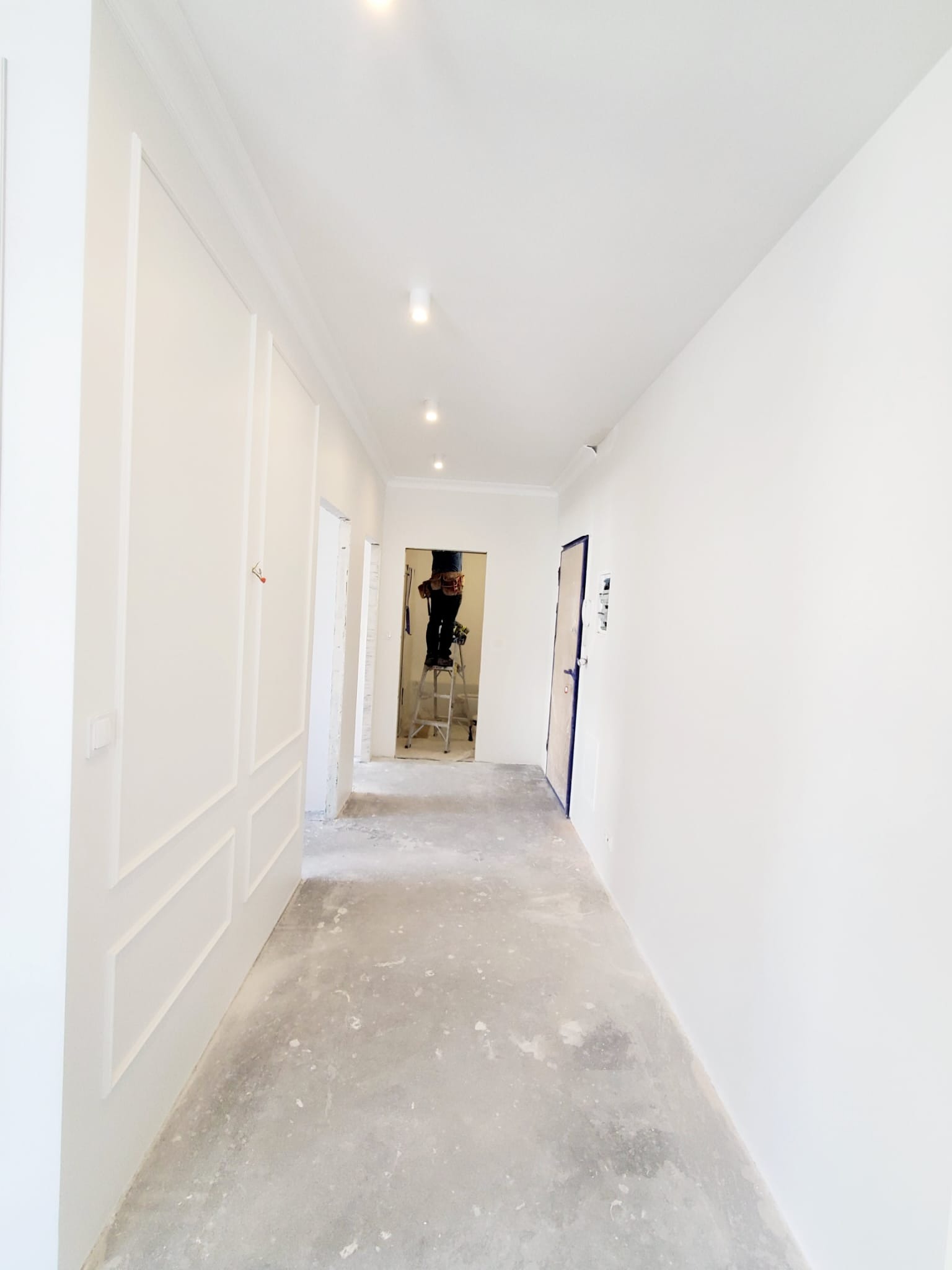 Korytarz w trakcie malowania na biało, widoczny pracownik na drabinie, świeżo pomalowane ściany z dekoracyjnymi panelami i sztukaterią, surowa podłoga zabezpieczona folią.