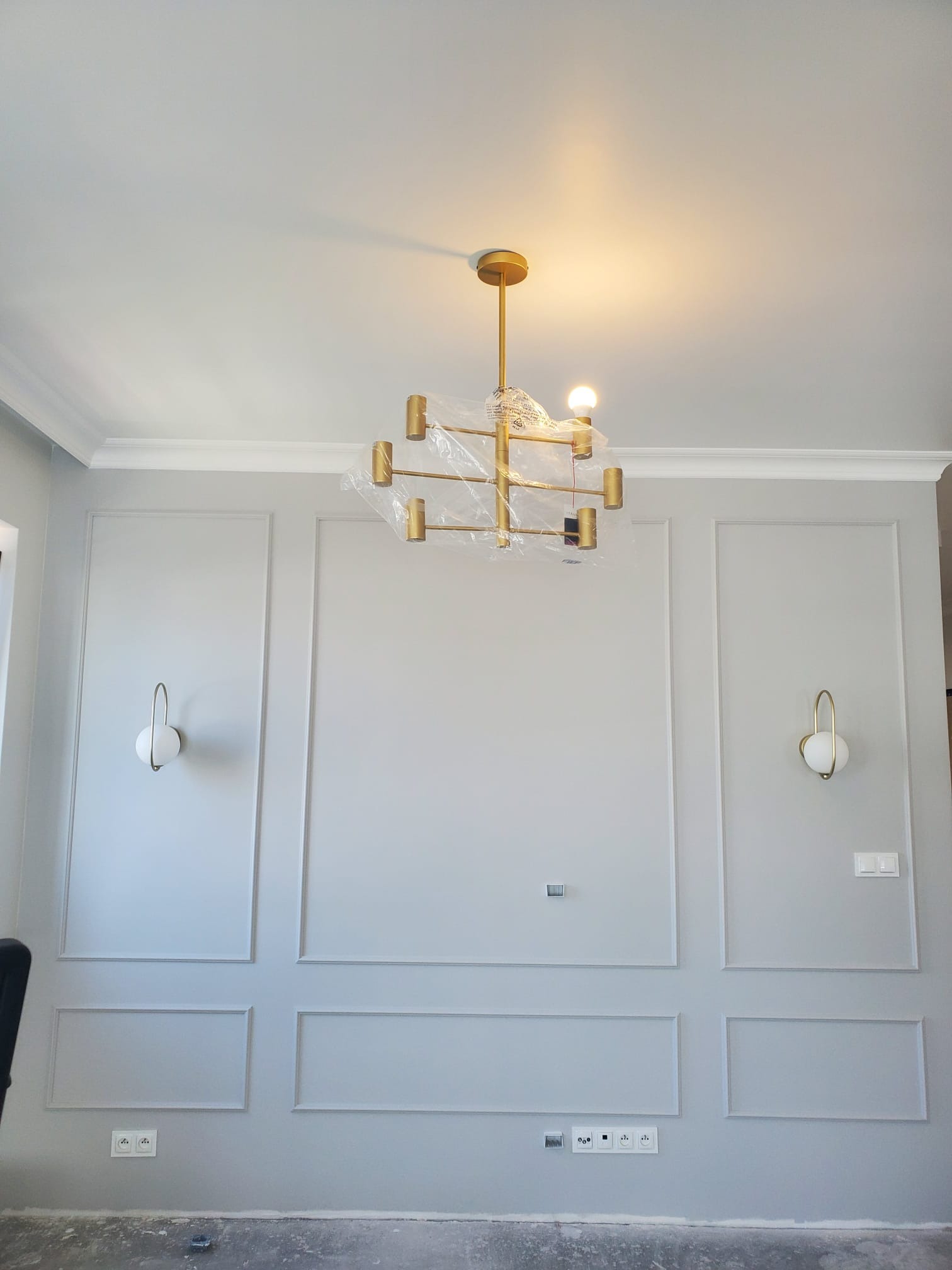 Szara ściana z dekoracyjnymi listwami, dwoma kinkietami z kulistymi kloszami i złotym, nowoczesnym żyrandolem w folii ochronnej, widoczne gniazdka elektryczne.
