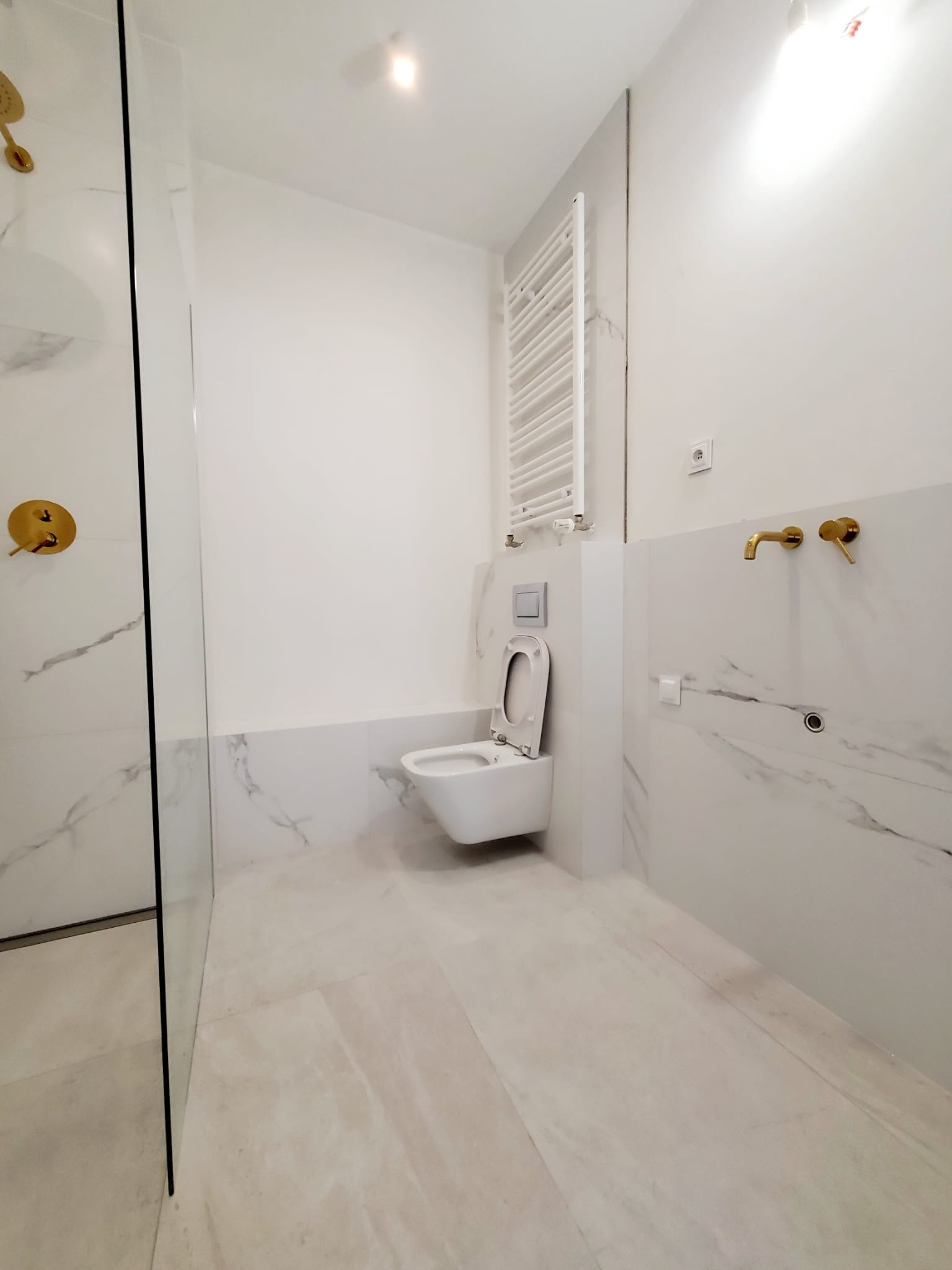 Nowoczesna łazienka w bieli z elementami marmuru, złotą armaturą i grzejnikiem drabinkowym.