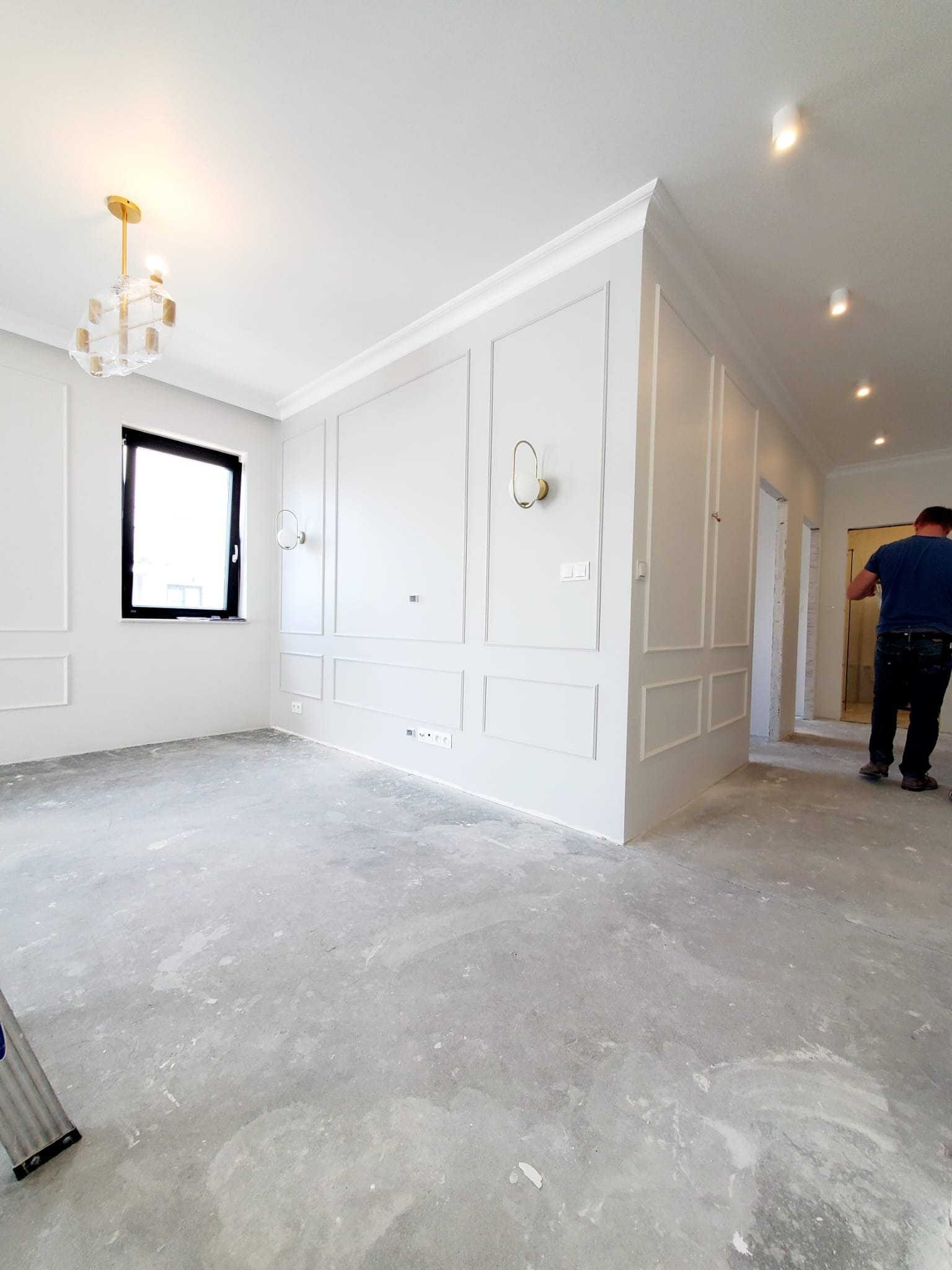 Pokój w trakcie wykańczania w stylu klasycznym: białe ściany z dekoracyjnymi panelami, nowoczesne oświetlenie, surowy beton na podłodze, widoczny pracownik w tle.