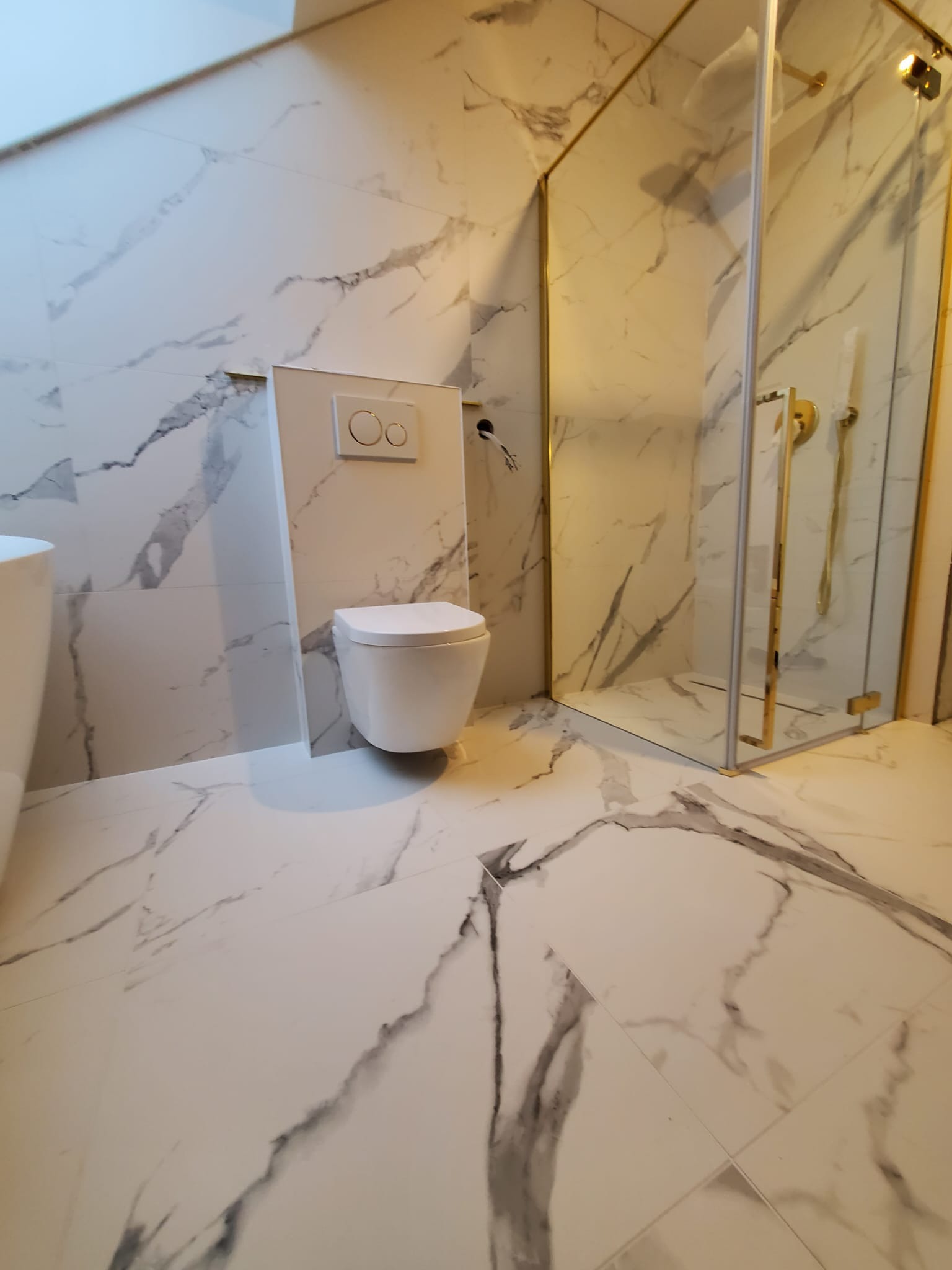 Nowoczesna łazienka z białymi płytkami imitującymi marmur, wisząca toaleta z podtynkową spłuczką oraz kabina prysznicowa ze złotymi elementami wykończeniowymi.