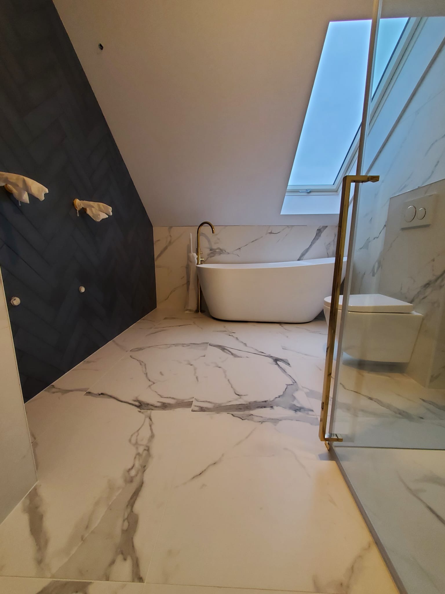 Nowoczesna łazienka na poddaszu z marmurową podłogą i ścianami, wolnostojąca wanna z złotą armaturą, toaleta podwieszana i kabina prysznicowa ze złotymi detalami.