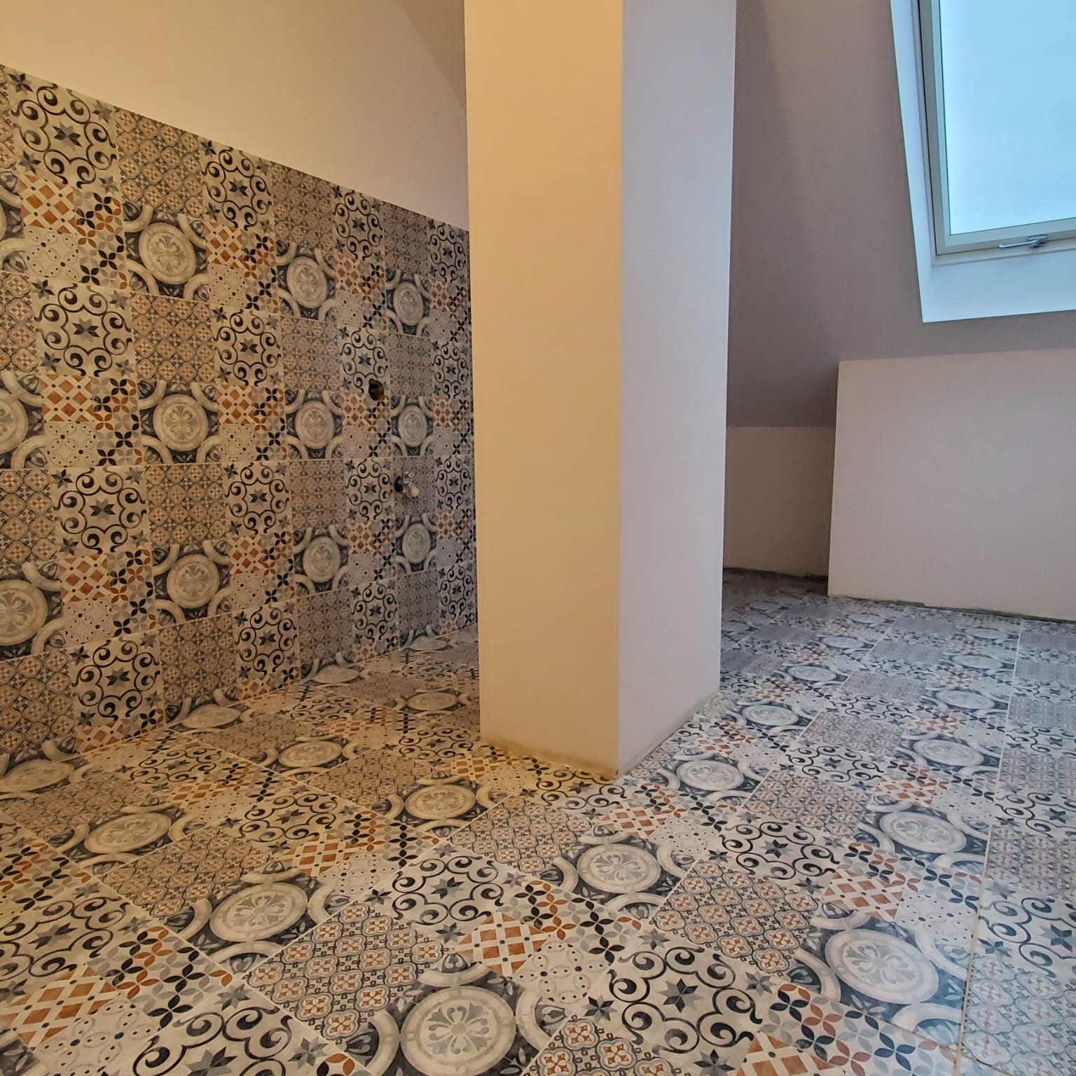 Ukończona łazienka na poddaszu z wzorzystymi płytkami na podłodze i ścianach, widoczny pionowy słup i okno dachowe.