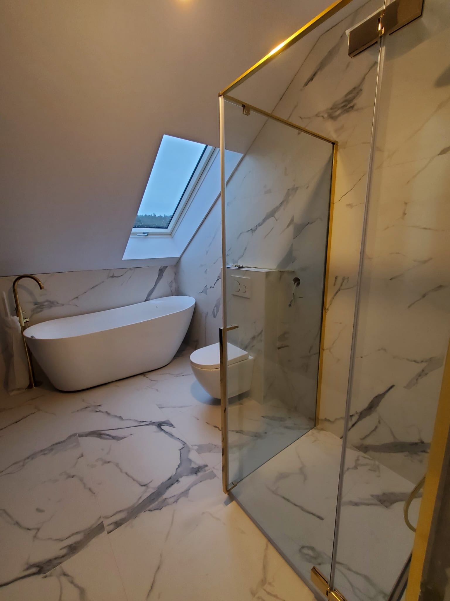 Elegancka łazienka w Warszawie na poddaszu z marmurowymi płytkami, wolnostojącą wanną, toaletą podtynkową i kabiną prysznicową z elementami w kolorze złotym.
