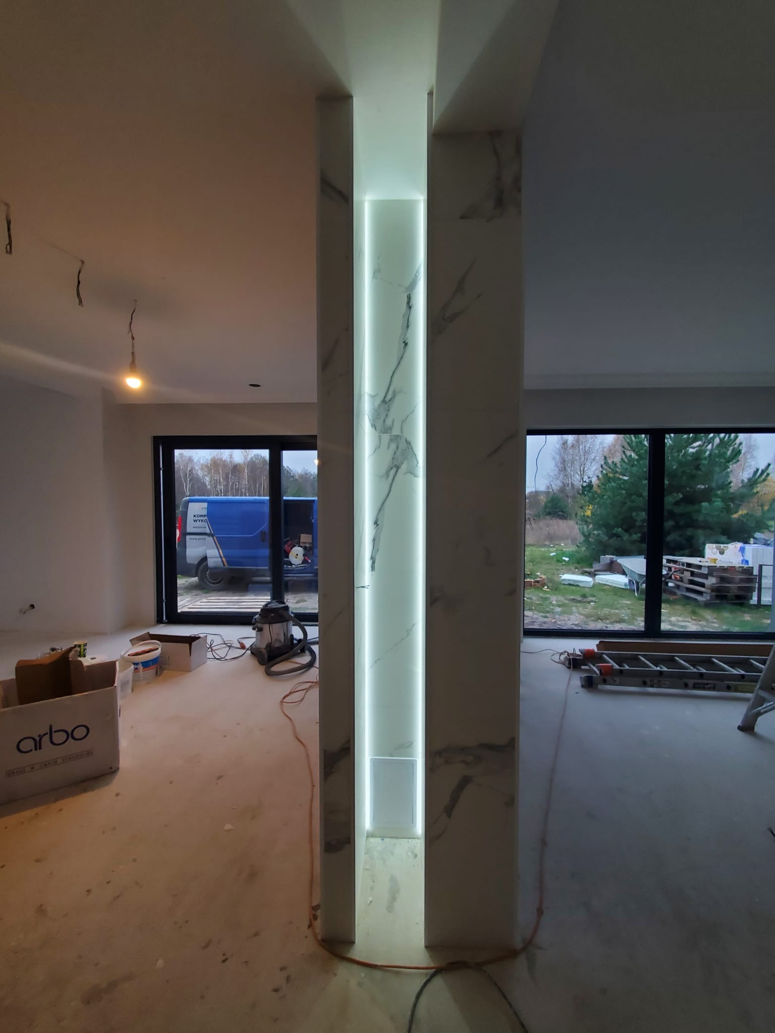 Wnętrze w trakcie prac wykończeniowych: kolumna z podświetleniem LED imitująca marmur, widoczne narzędzia i materiały budowlane, okno z widokiem na zewnątrz