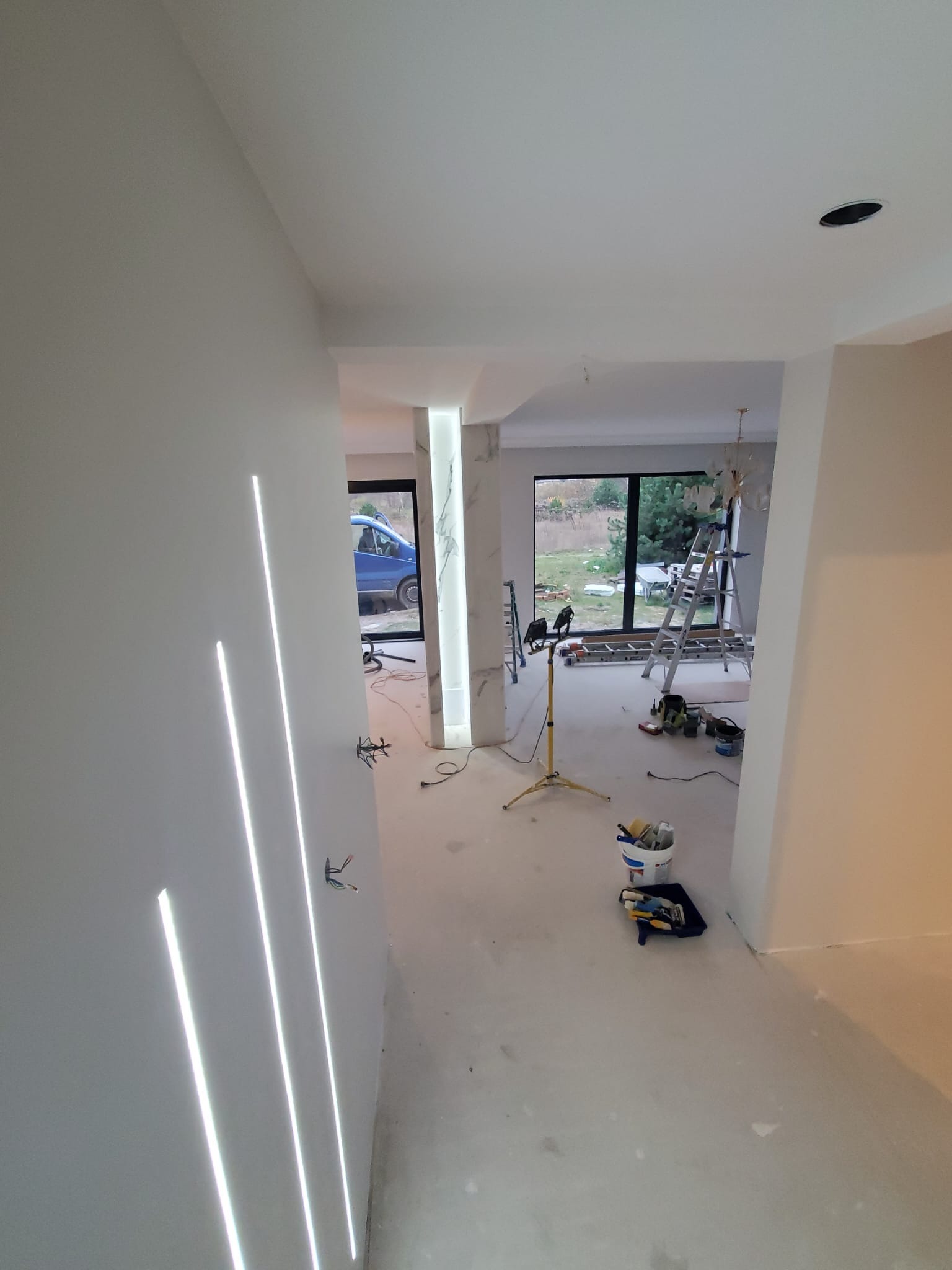 Wnętrze w trakcie prac wykończeniowych: ściany pomalowane na biało, wbudowane pionowe oświetlenie LED, widoczne narzędzia malarskie, drabina i lampa budowlana, okno z widokiem na ogród.