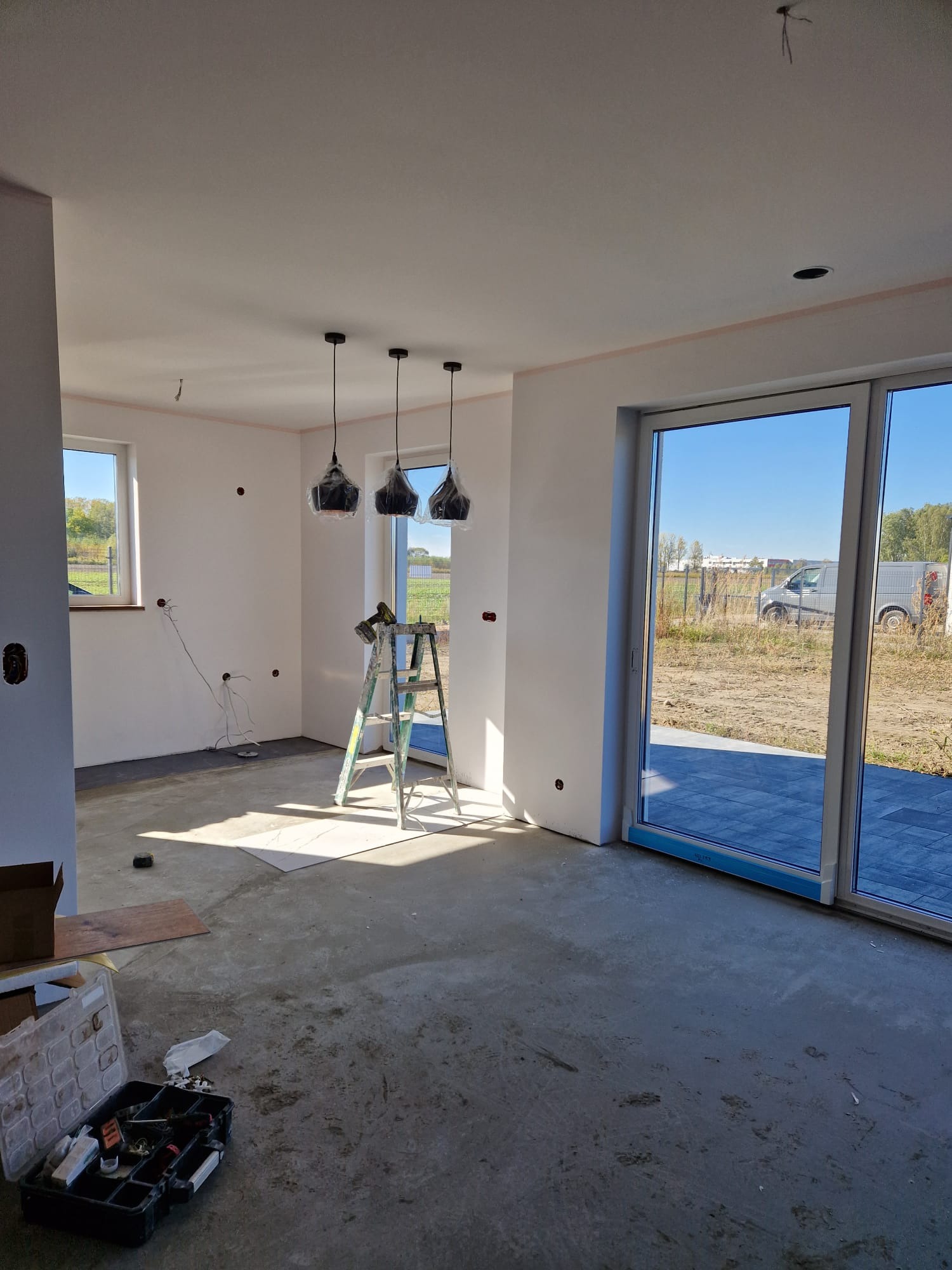 Wnętrze domu w trakcie prac wykończeniowych: widoczne ściany po malowaniu, surowe podłoże, drabina, wiszące lampy w folii ochronnej, skrzynka z narzędziami i przesuwne drzwi tarasowe z widokiem...
