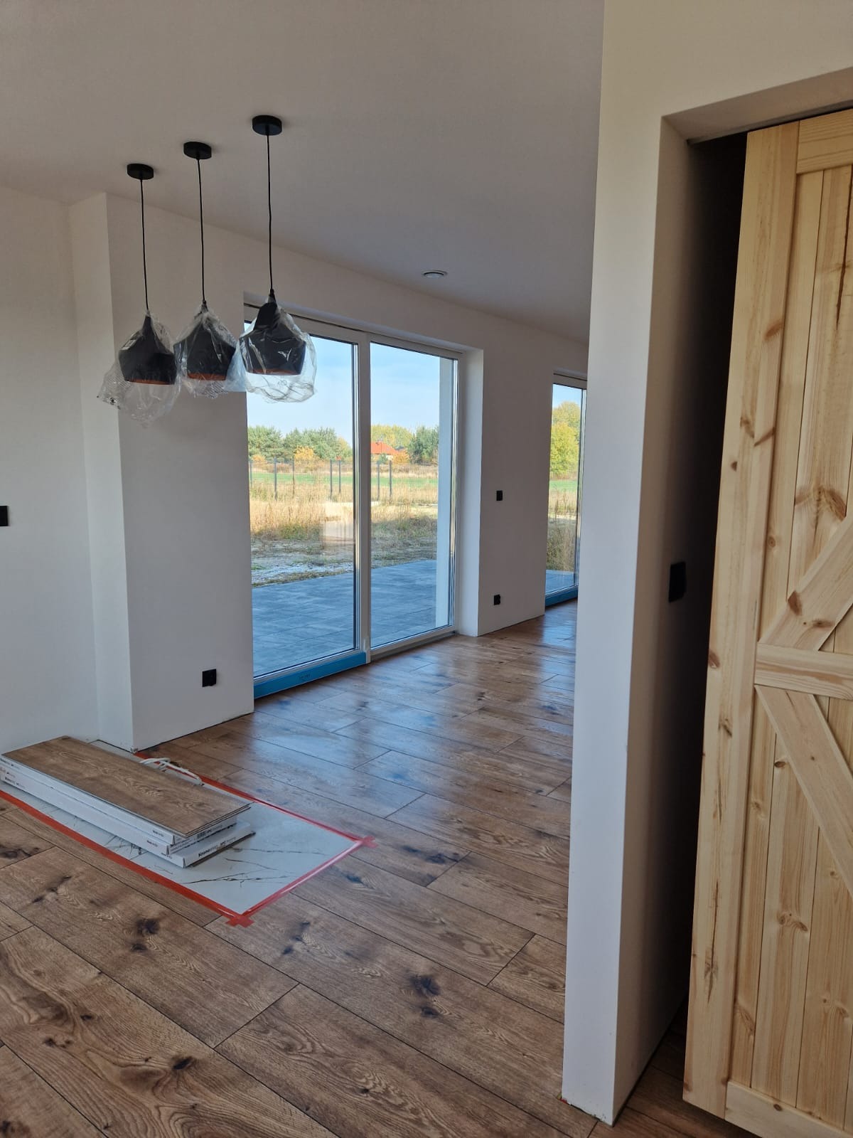 Wnętrze domu w trakcie wykańczania: drewniana podłoga, przesuwne drzwi stodołowe, lampy wiszące w folii ochronnej, okna tarasowe z widokiem na ogród, panele podłogowe w oryginalnym opakowaniu.