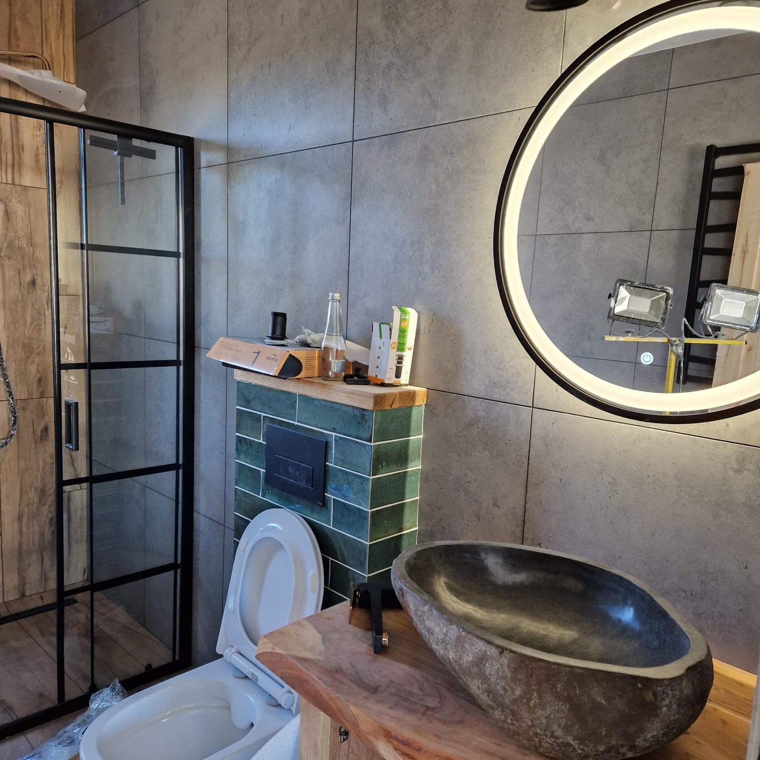 Nowoczesna łazienka z kabiną prysznicową z czarnymi profilami, umywalką z kamienia na drewnianym blacie i okrągłym lustrem z podświetleniem LED, szare płytki na ścianach.