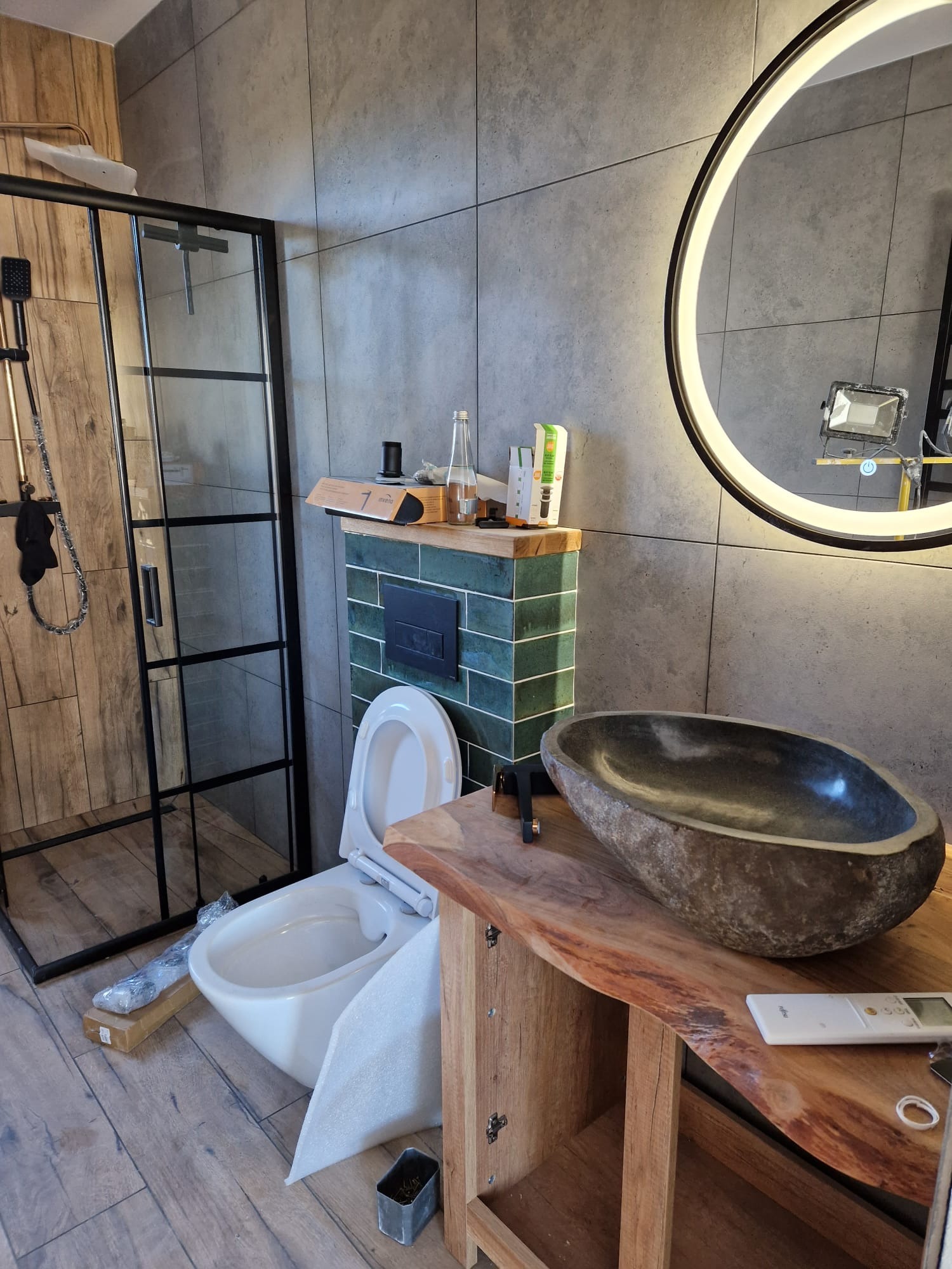Nowoczesna łazienka w trakcie wykańczania: kabina prysznicowa z czarną ramą, misa umywalkowa z kamienia na blacie z surowego drewna, podświetlane okrągłe lustro, wisząca toaleta z otwartą klapą...