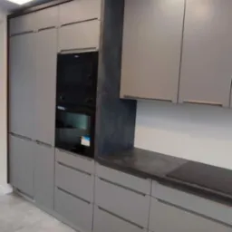 Szara kuchnia na wymiar z zabudowaną piekarnikiem i płytą indukcyjną na blacie z ciemnego kamienia, oświetlenie punktowe sufitowe.