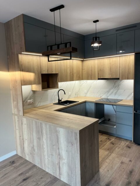 Nowoczesna kuchnia z wyspą, fronty szafek w odcieniach szarości i drewna, podświetlenie LED pod szafkami górnymi, czarny zlew i armatura, marmurowy backsplash.