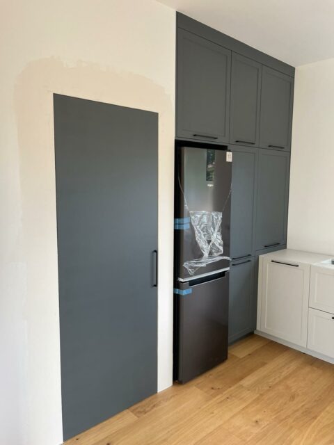 Nowoczesna kuchnia z szarymi szafkami na wymiar, ukrytymi drzwiami i nową lodówką w folii ochronnej, widoczne drewniane panele na podłodze.