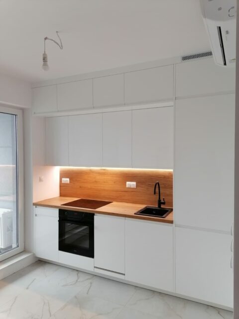 Nowoczesna, minimalistyczna kuchnia z białymi frontami, drewnianym blatem i czarną armaturą, oświetlenie LED pod szafkami, marmurowe płytki na podłodze, widoczny fragment okna i klimatyzator.