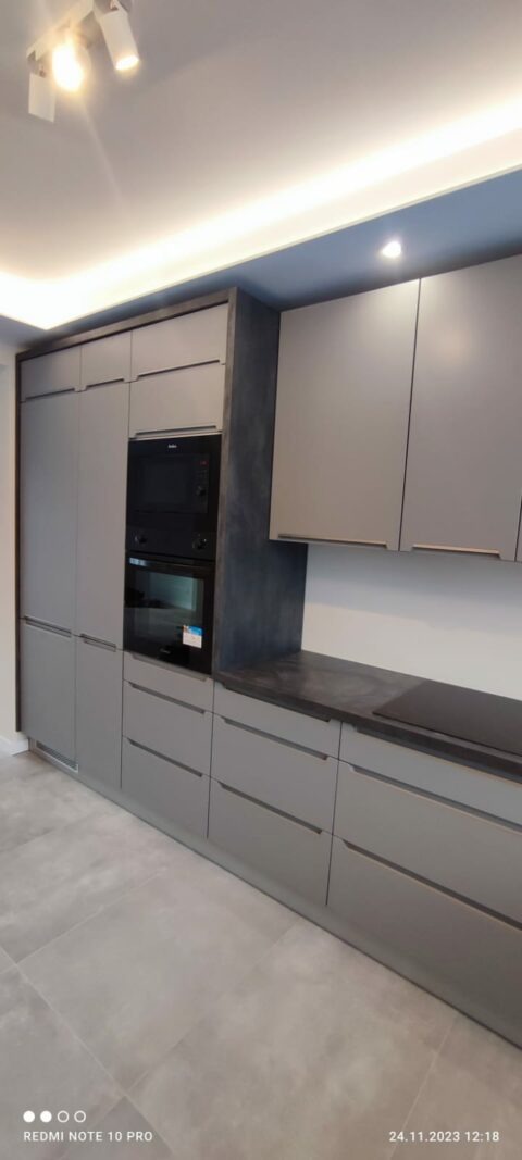 Szara kuchnia na wymiar z zabudowaną piekarnikiem i płytą indukcyjną na blacie z ciemnego kamienia, oświetlenie punktowe sufitowe.