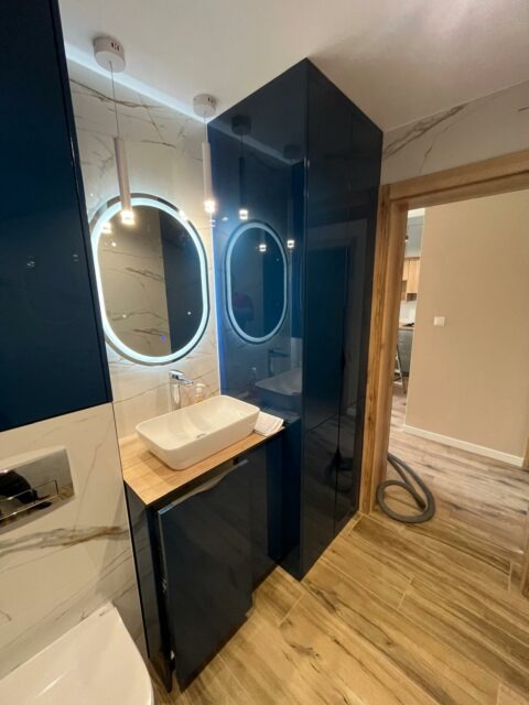 Nowoczesna łazienka z szafką pod umywalkę w kolorze granatowym, drewnianym blatem i owalnym lustrem z podświetleniem LED, obok wysoka, lakierowana szafa wnękowa.