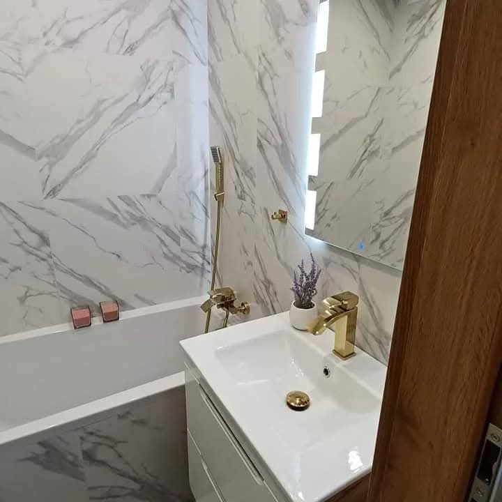 Wykończona łazienka z wanną obłożoną marmurową płytką, złota armatura, umywalka z szafką i podświetlane lustro, dekoracyjna lawenda w doniczce.