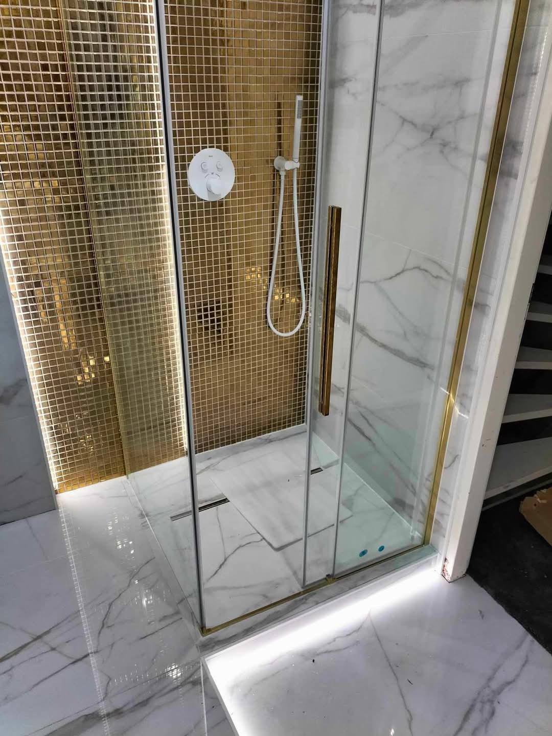 Kabina prysznicowa z złotą mozaiką na ścianie, białe marmurowe płytki na podłodze, oświetlenie LED w listwach przypodłogowych.