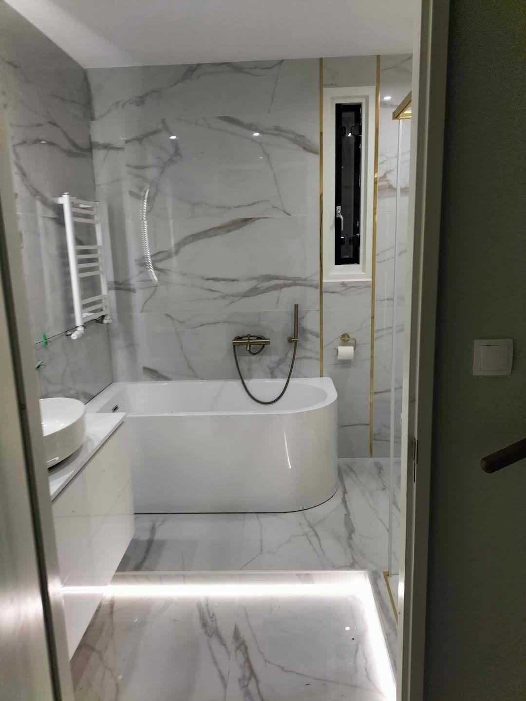 Nowoczesna łazienka z wanną, umywalką nablatową i marmurowymi płytkami z podświetleniem LED wzdłuż podłogi, akcenty w kolorze złotym.