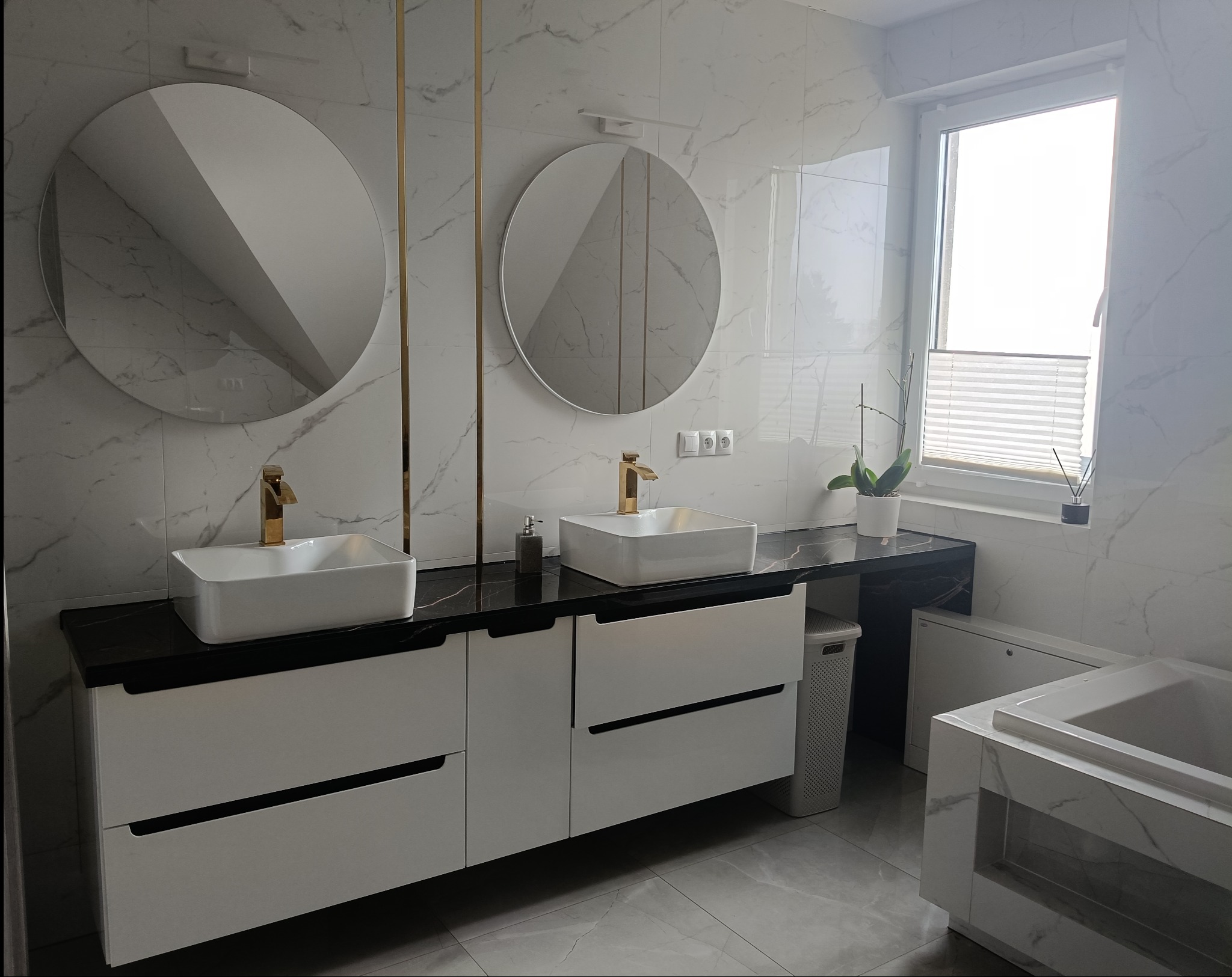 Nowoczesna łazienka z marmurowymi płytkami, dwoma okrągłymi lustrami z złotymi akcentami, białymi szafkami z czarnymi blatami i złotymi bateriami, oraz białą wanną.
