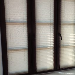 PAMI SPÓŁKA Z OGRANICZONĄ ODPOWIEDZIALNOŚCIĄ - Beżowe, poziome przesłony okienne zamontowane w oknie z ciemnobrązowymi ramami, widoczne uchwyty okienne.