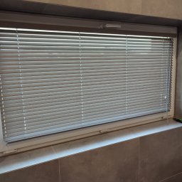 PAMI SPÓŁKA Z OGRANICZONĄ ODPOWIEDZIALNOŚCIĄ - Zamknięte, poziome, szare listwy w oknie z widocznymi sznurkami regulującymi, w tle szare płytki ścienne.