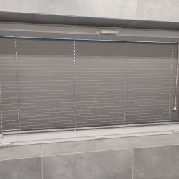 PAMI SPÓŁKA Z OGRANICZONĄ ODPOWIEDZIALNOŚCIĄ - Zamknięte szare żaluzje aluminiowe w oknie, widoczne z bliska, z fragmentem parapetu wyłożonego szarymi płytkami.
