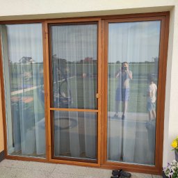 PAMI SPÓŁKA Z OGRANICZONĄ ODPOWIEDZIALNOŚCIĄ - Brązowe okno tarasowe z zamontowaną siatką przeciw owadom, widoczne odbicie osoby robiącej zdjęcie i dziecka, na podłodze leżą buty.