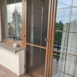 PAMI SPÓŁKA Z OGRANICZONĄ ODPOWIEDZIALNOŚCIĄ - Brązowa moskitiera ramkowa zamontowana w drzwiach balkonowych, widok z zewnątrz, na parapecie stoi kubek.