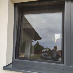 PAMI SPÓŁKA Z OGRANICZONĄ ODPOWIEDZIALNOŚCIĄ - Okna z zamontowaną siatką przeciw owadom, widoczna faktura siatki oraz odbicie otoczenia i osoby robiącej zdjęcie.