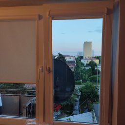 PAMI SPÓŁKA Z OGRANICZONĄ ODPOWIEDZIALNOŚCIĄ - Okno z częściowo opuszczoną roletą materiałową w kolorze jasnoszarym, widok na osiedle zza okna, fragment zasłony po prawej stronie.