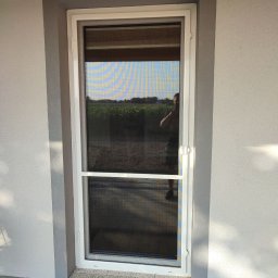 PAMI SPÓŁKA Z OGRANICZONĄ ODPOWIEDZIALNOŚCIĄ - Zamknięta, biała moskitiera ramkowa na drzwi balkonowe, z widocznym odbiciem osoby robiącej zdjęcie na tle zielonego pola.