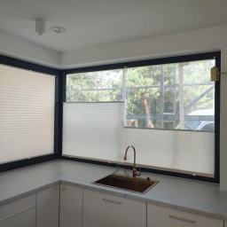 PAMI SPÓŁKA Z OGRANICZONĄ ODPOWIEDZIALNOŚCIĄ - Jasna kuchnia z oknami wyposażonymi w białe, plisowane osłony okienne, widok na zewnątrz zza drzew.