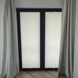 PAMI SPÓŁKA Z OGRANICZONĄ ODPOWIEDZIALNOŚCIĄ - Okno z zamontowanymi plisami w kolorze białym, zasłonięte szarymi zasłonami, na podłodze parkiet ułożony w jodełkę.