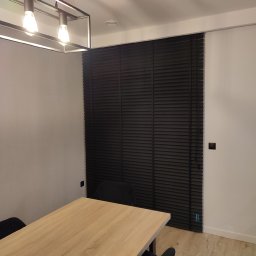 PAMI SPÓŁKA Z OGRANICZONĄ ODPOWIEDZIALNOŚCIĄ - Ciemne żaluzje drewniane w minimalistycznym wnętrzu nad stołem z jasnego drewna, oświetlone nowoczesną lampą.