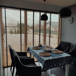 PAMI SPÓŁKA Z OGRANICZONĄ ODPOWIEDZIALNOŚCIĄ - Jadalnia z drewnianymi poziomymi przesłonami okiennymi w kolorze jasnego drewna, z czarnymi listwami, widoczne przez okno zasypane śniegiem podwórko i domy.