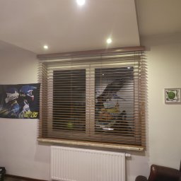 PAMI SPÓŁKA Z OGRANICZONĄ ODPOWIEDZIALNOŚCIĄ - Zamknięte żaluzje drewniane w oknie, z widocznym plakatem motorowym po lewej stronie i grzejnikiem pod parapetem.