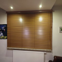 PAMI SPÓŁKA Z OGRANICZONĄ ODPOWIEDZIALNOŚCIĄ - Zamknięte poziome osłonki okienne w kolorze drewna, zamontowane nad grzejnikiem, z plakatem na ścianie obok.