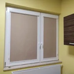 Dwa okna PCV z beżowymi roletami zwijanymi, zamontowane w żółtej ścianie nad grzejnikiem.