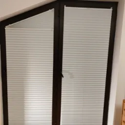 Białe plisy zamontowane w oknie dachowym o nieregularnym kształcie, widoczna drewniana podłoga i fragment komody.