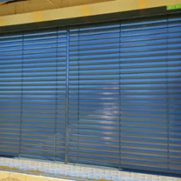 Zamknięte, poziome lamele w oknie budynku w trakcie budowy, widoczne bloczki betonowe i elementy montażowe.