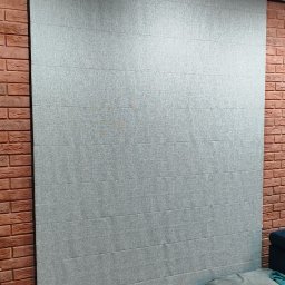 PAMI SPÓŁKA Z OGRANICZONĄ ODPOWIEDZIALNOŚCIĄ - Szara, poziomo składana osłona okienna zamontowana na ścianie z czerwonej cegły, oświetlona reflektorami sufitowymi.