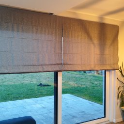 PAMI SPÓŁKA Z OGRANICZONĄ ODPOWIEDZIALNOŚCIĄ - Szara, tekstylna roleta w stylu rzymskim, zamontowana w nowoczesnym salonie z widokiem na trawnik.