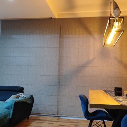 PAMI SPÓŁKA Z OGRANICZONĄ ODPOWIEDZIALNOŚCIĄ - Jasnoszara tkanina zasłaniająca okno, tworząca poziome pasy, widoczna w salonie z drewnianą podłogą i elementami wystroju, takimi jak lampa z ozdobną figurką.