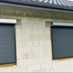 Dwa okna z zamontowanymi, częściowo opuszczonymi, szarymi roletami zewnętrznymi na surowej, białej ścianie budynku w trakcie budowy, z widoczną pianką montażową wokół okien.