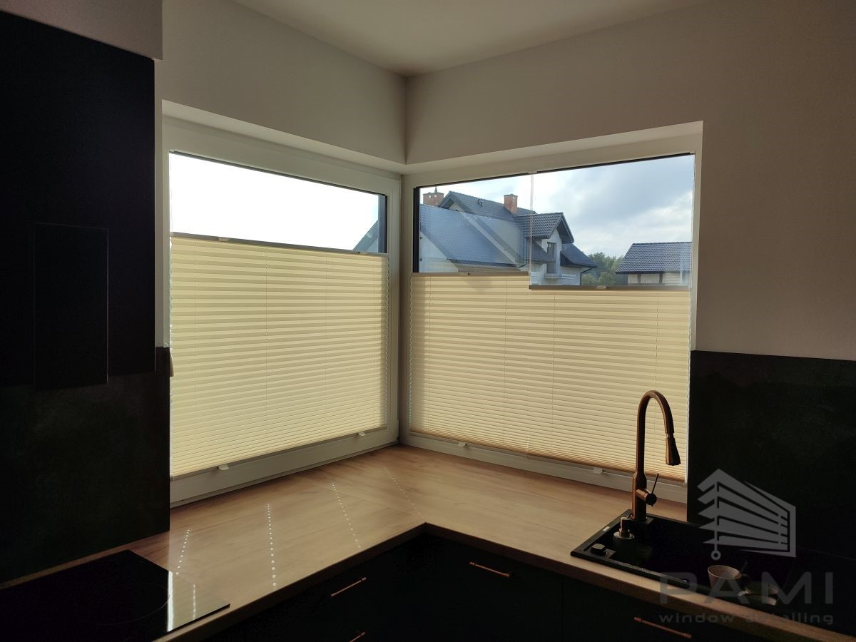Beżowe, harmonijkowe osłony okienne zamontowane w narożnym oknie kuchennym z widokiem na domy. Widoczna bateria kuchenna w kolorze miedzi i fragment blatu.